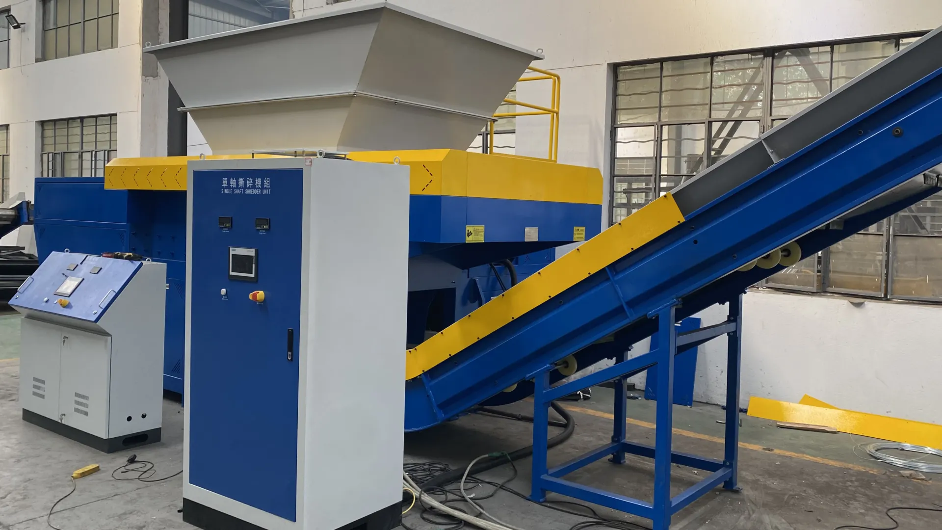 An industrial facility housing a rigid plastics shredder. The shredder features a robust blue and yellow design with a large white feed hopper on top. A blue and yellow conveyor belt system is used for material input. The shredder is equipped with control panels for operation and monitoring, emphasizing ease of use and efficiency. This setup is designed to handle and process rigid plastic materials effectively, making it suitable for recycling and waste management applications. The overall design highlights the machine’s durability and high performance in processing various types of rigid plastics.