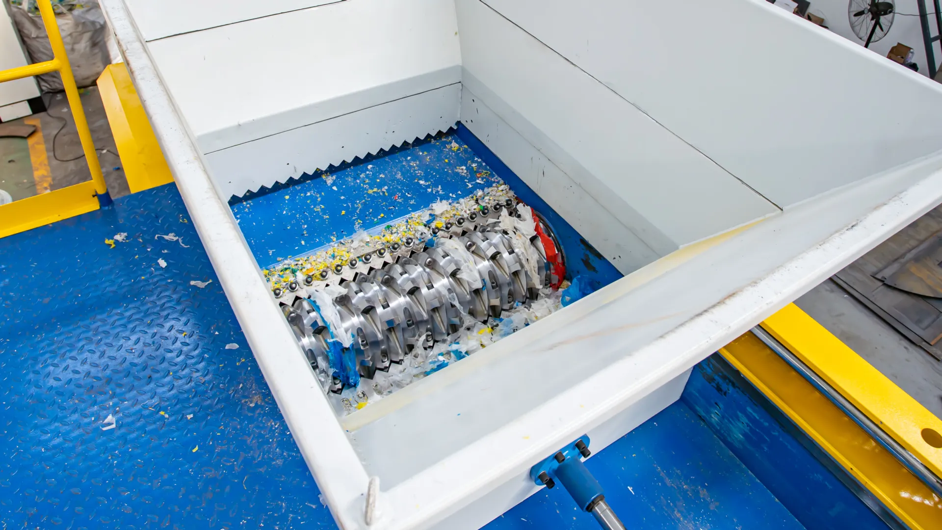 Een close-up van het interne snijmechanisme van een shredder met één as. De afbeelding toont de robuuste messen en tanden van de shredder, ontworpen om verschillende materialen af te breken. De snijkamer heeft een blauwe basis en witte wanden, waarbij versnipperd materiaal zichtbaar is rond de messen. De machine is ontworpen voor een hoge efficiëntie en duurzaamheid bij het verwerken van harde kunststoffen en andere materialen. De omringende gele en blauwe structuur benadrukt de industriële setting en benadrukt de robuuste constructie en functionaliteit van de machine.