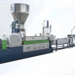 Imaginea prezintă o linie industrială de extrudare a plasticului. Acest utilaj este utilizat pentru producția de produse din plastic prin topirea materiei prime din plastic și formarea acesteia într-un profil continuu. Echipamentul include diverse componente, cum ar fi un buncăr pentru intrarea materiei prime, un extruder în care plasticul este topit și împins printr-o filieră pentru a-l forma, sisteme de răcire și, eventual, mecanisme de tăiere sau de laminare pentru manipularea produsului final. Instalația este utilizată de obicei în procesele de fabricație pentru crearea de țevi, foi, filme și alte profile din plastic.