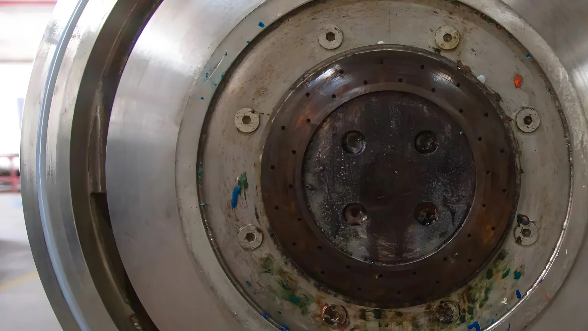 The picture shows a close-up view of the interior part of a pelletizer used for processing PP (polypropylene) and PE (polyethylene) films into plastic granules. This part of the machinery appears to be the die face where molten plastic is extruded and cut into pellets. You can see remnants of plastic materials in various colors clinging to the metal surfaces, indicating recent use or the ongoing processing of recycled plastics. The large circular metal surface with holes and grooves is typical of die face cutters in plastic recycling operations, which help form and cut the extruded plastic into uniform pellets.