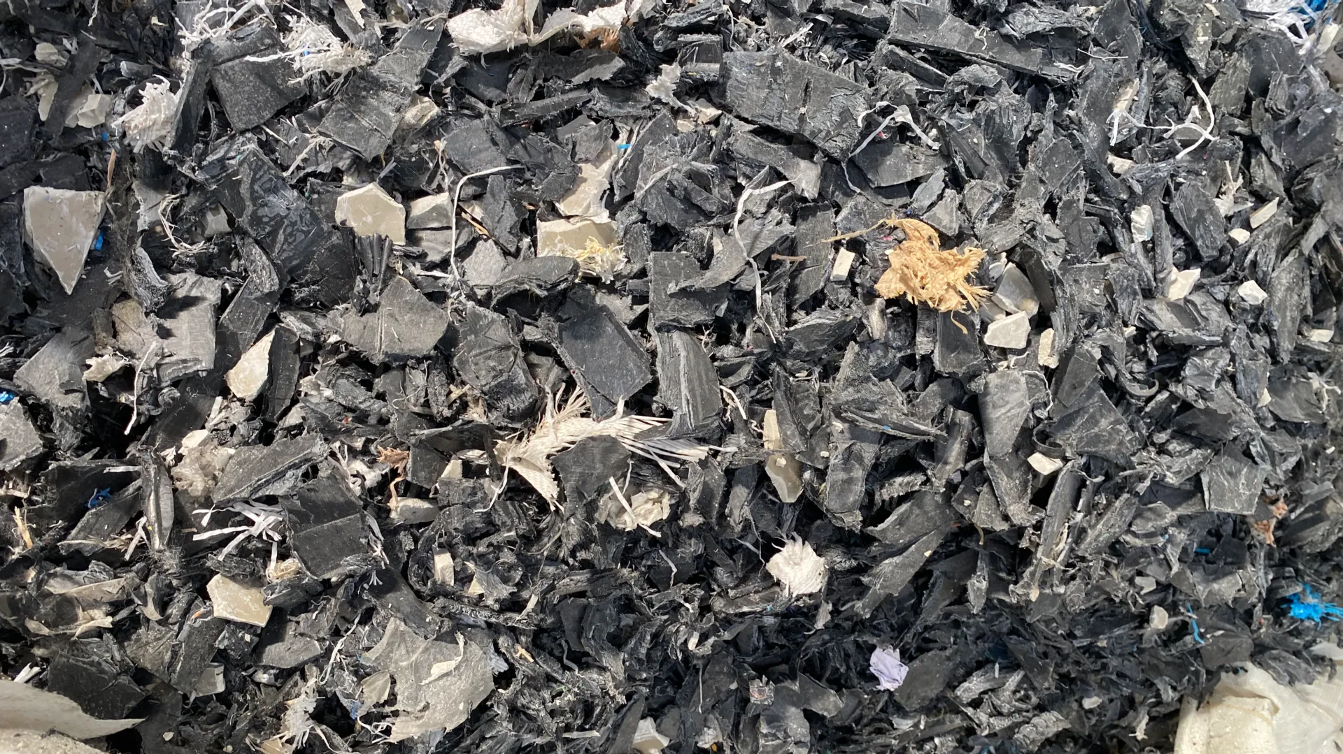 Close-up view of shredded plastic waste, showcasing a mix of black, gray, and white plastic fragments. The image highlights the effectiveness of industrial shredders in breaking down plastic materials into smaller, manageable pieces for recycling. The shredded pieces vary in size and shape, demonstrating the shredder’s ability to handle diverse types of plastic waste. This is a crucial step in the plastic recycling process, enabling further processing and reuse of materials.