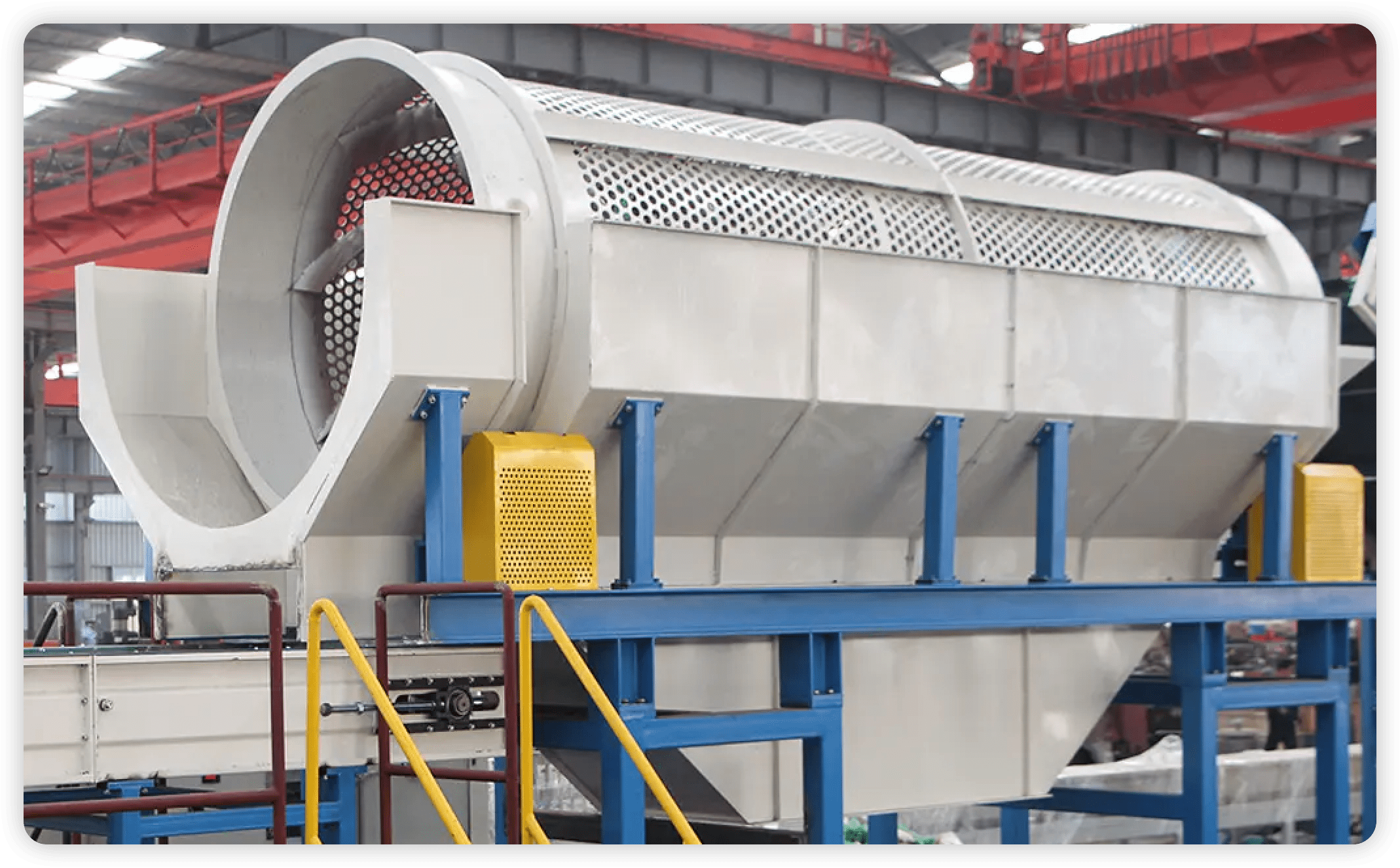 An industrial rotary drum screen used for material sorting and separation. The drum screen, made of perforated metal, is mounted on a sturdy blue and yellow frame. It features a cylindrical shape and is slightly inclined for efficient material movement. The machine operates within an industrial facility, designed to filter and separate different sizes of materials during the recycling or waste processing stages. The equipment is equipped with safety railings and protective covers to ensure safe operation.