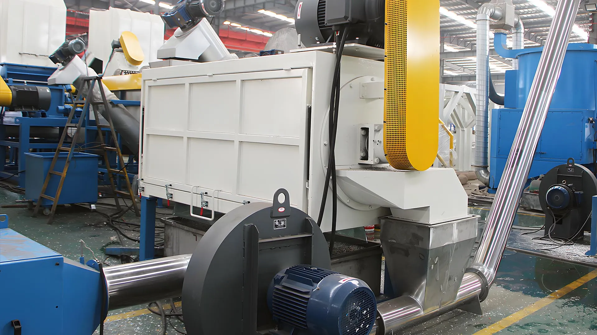 Industrial-grade high-speed friction washer operating inside a plastic recycling facility, featuring large yellow and blue machinery with metal conduit piping and electric motors. The setting is clean and well-organized, highlighting the efficiency and automation of modern recycling technologies.