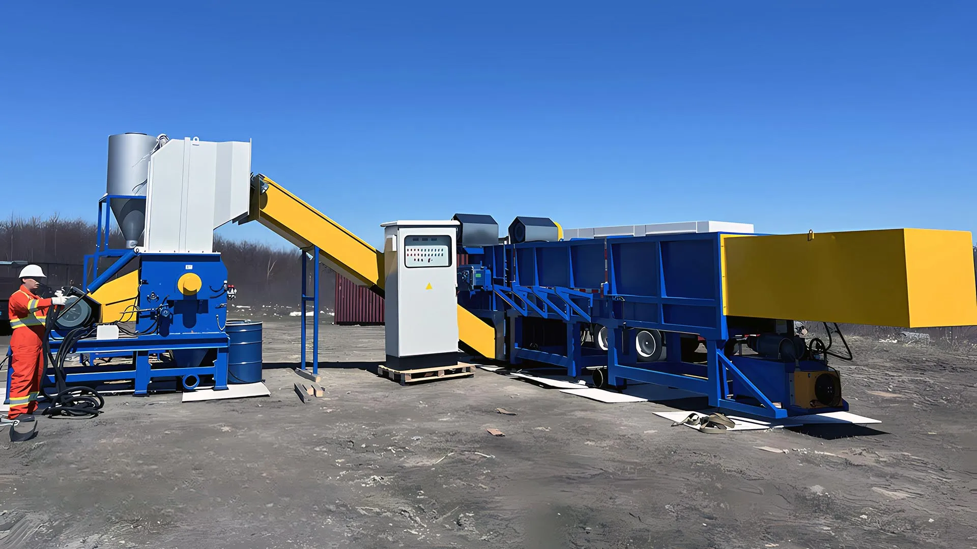 a mobile industrial shredding system for pipes, set in a barren outdoor area. The system is primarily composed of several interconnected units painted in vibrant blue and yellow, enhancing visibility. On the left, a worker in a bright orange suit and white helmet is operating the machinery, which includes a loading platform, a shredding unit, and a sorting conveyor. The clear blue sky and the lack of vegetation in the background suggest a remote or industrial location.