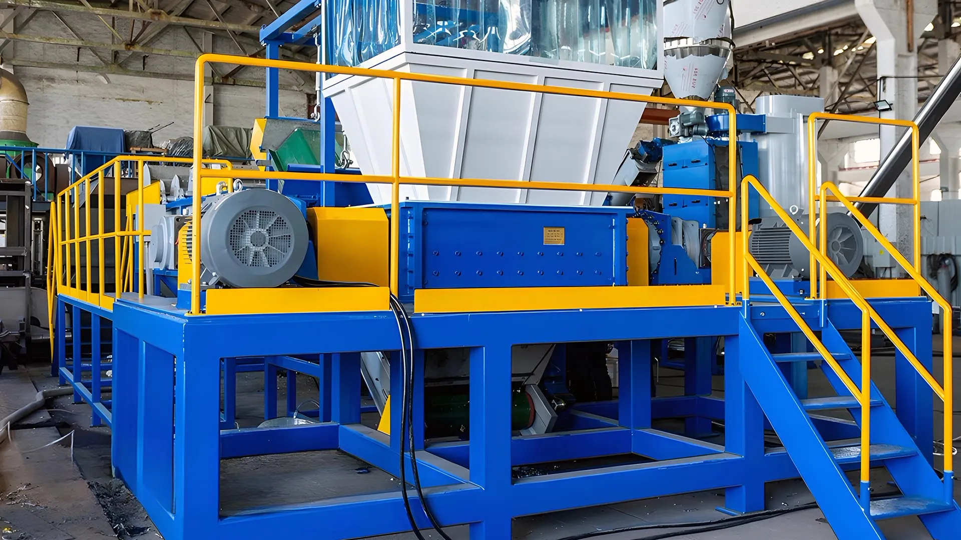 A robust recycling setup for shredding and washing rigid plastics such as PP, HDPE, and PS. The machinery, painted in vibrant blue and yellow, features a large hopper, shredding unit, and multiple conveyors within an industrial environment. Safety railings and steps provide secure access to operational areas, highlighting the facility’s commitment to worker safety and efficiency.