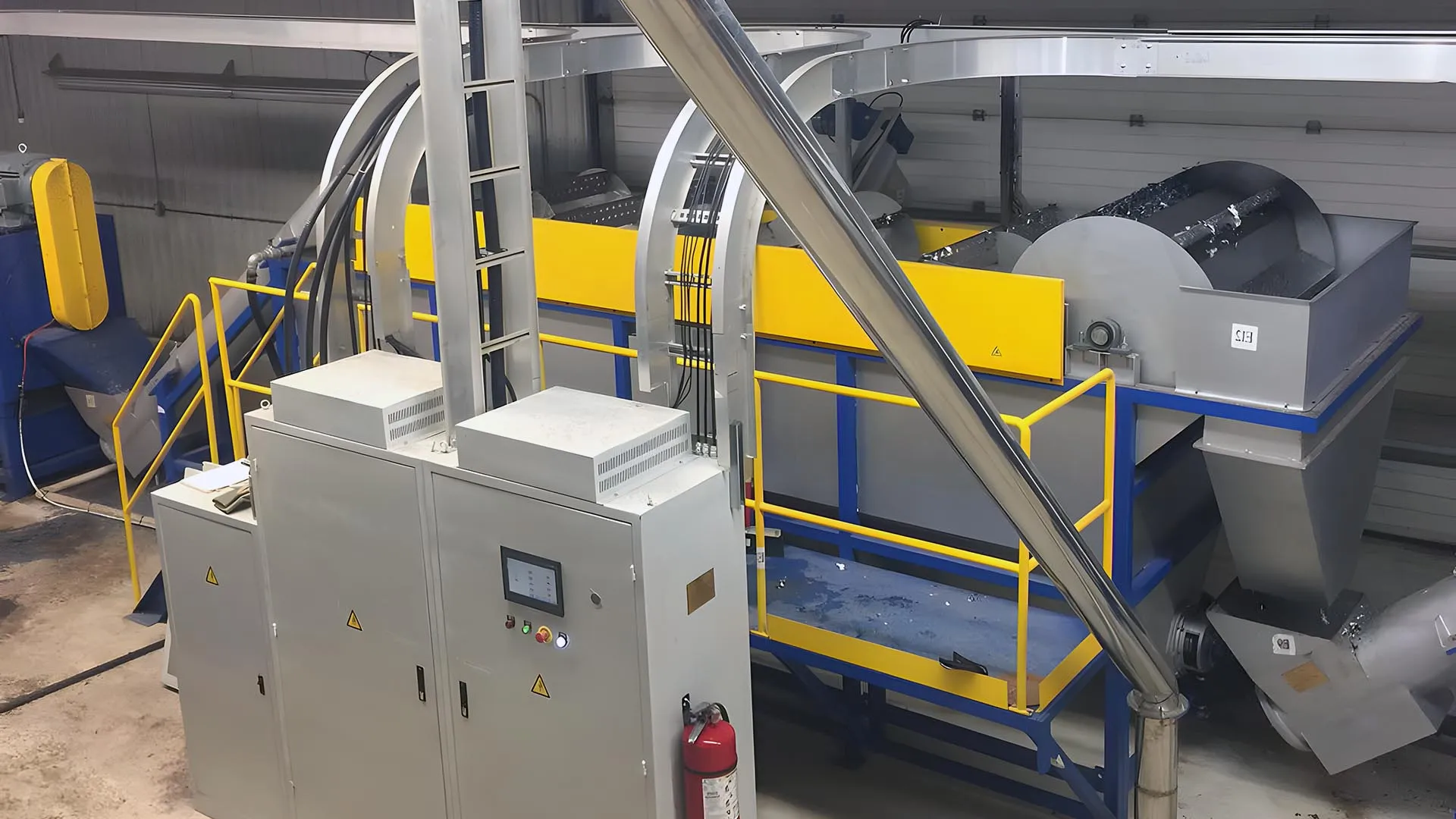 Interior of an industrial recycling facility featuring a PP and PE film regrind washing and recycling line. The equipment is painted in bright yellow and blue, enhancing visibility. It includes various conveyors, hoppers, and a cylindrical rotating drum, all integrated into a compact and efficient setup. Safety railings and an extinguisher are visible, emphasizing safety measures in place.